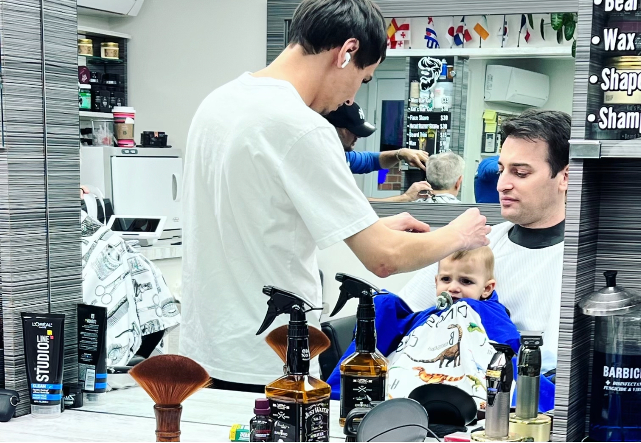 barber performing a hair cut for a toddler baby, baby sits on the lap of his father, so cute